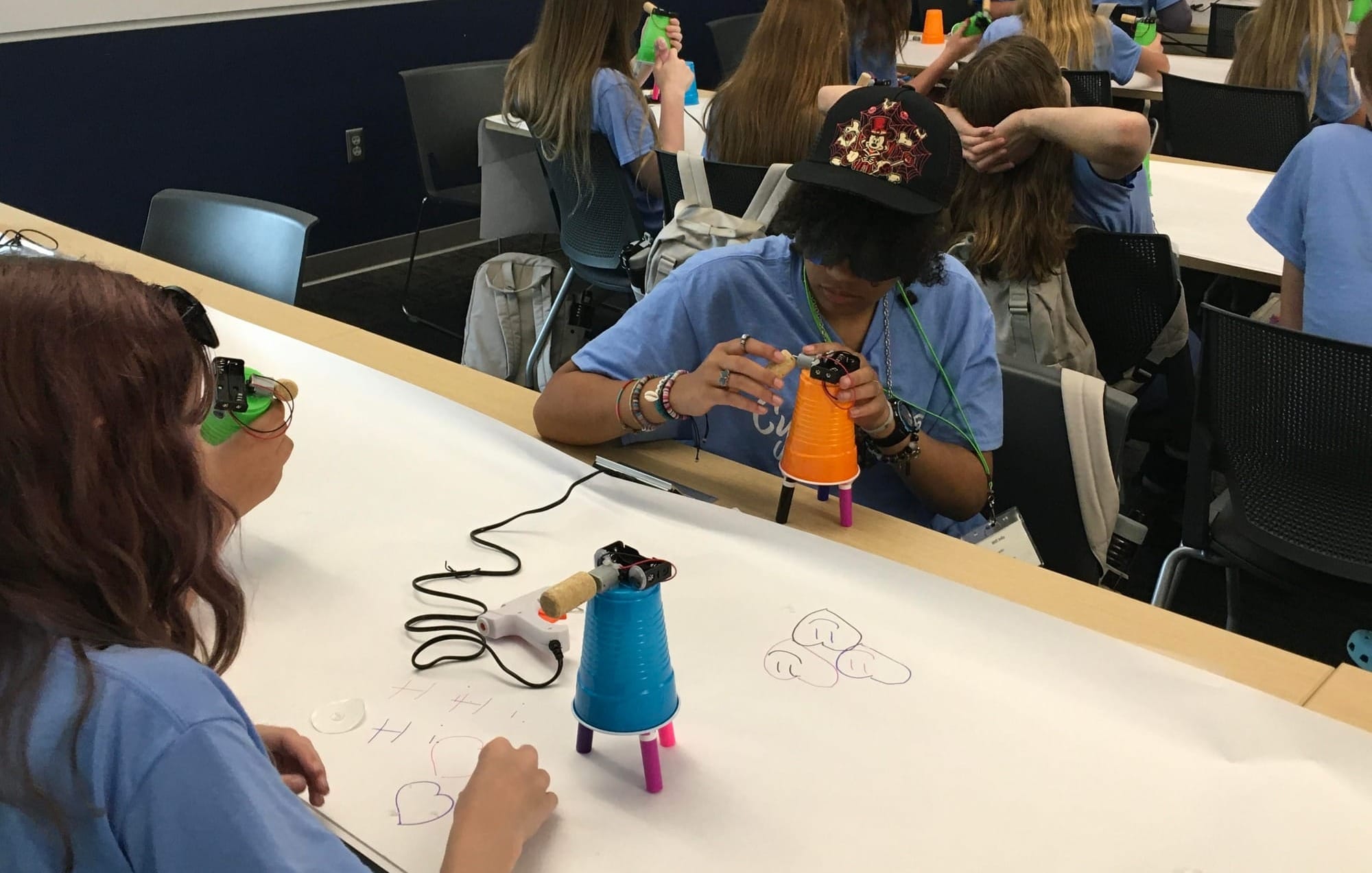 Girls at a table draw on a piece of paper using markers, a motor and a cup at the CybHER camp