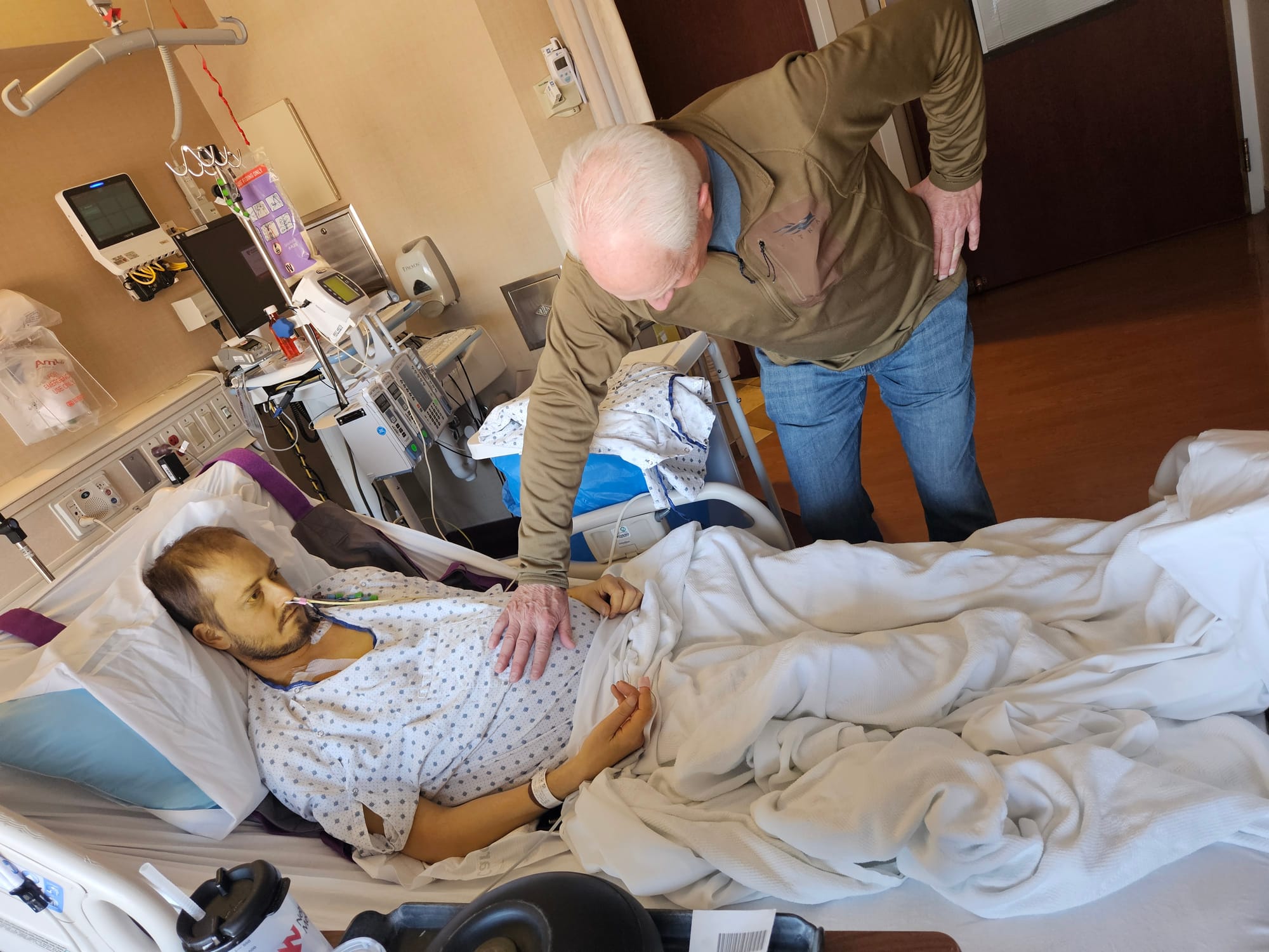 A man lays in a hospital bed with tubes coming out of his nose as his father touches his stomach.