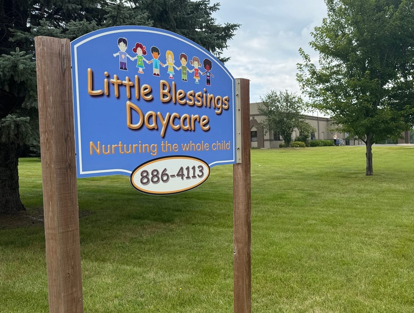 A blue Little Blessings Daycare sign sits in a grass field in front of a building