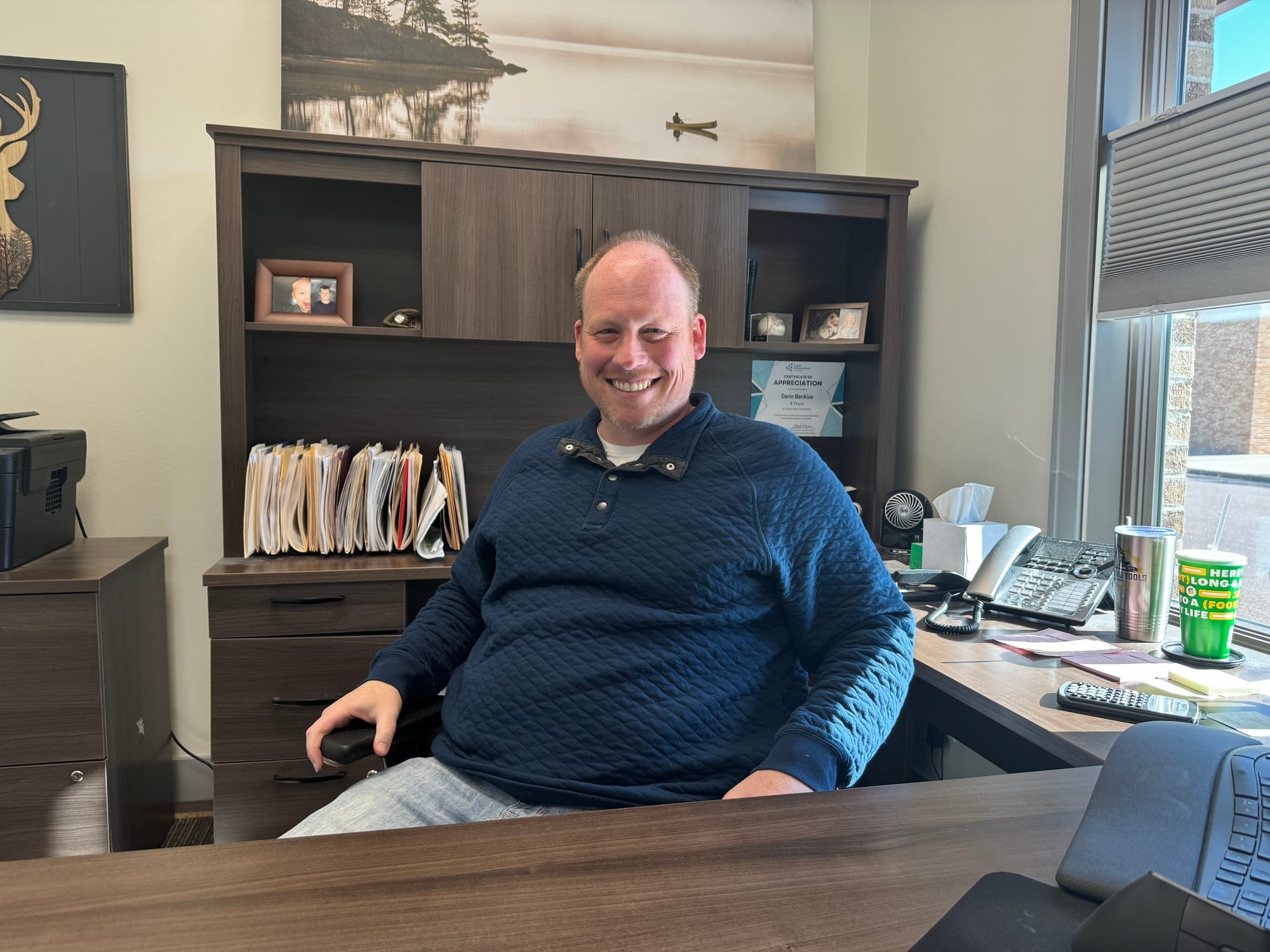 A man sits at behind his office desk and looks at the camera while smiling.