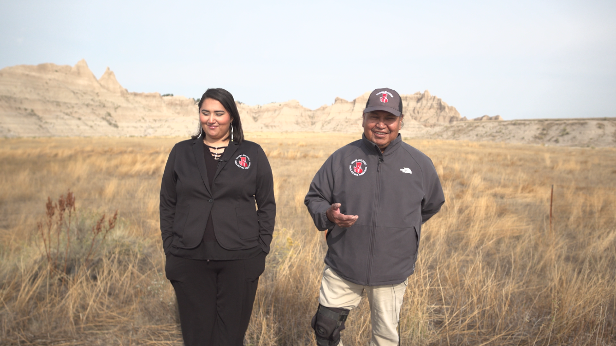 Tianna Yellow Hair (left) and her father, Guss Yellow Hair (right), walk through a prairie field with the Badlands of South Dakota in the background