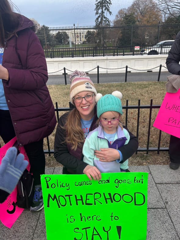 RiAnna Kolovsky and her daughter pose in front of the White House during a trip to Washington while holding a sign that says "policy comes and goes but Motherhood is here to stay"