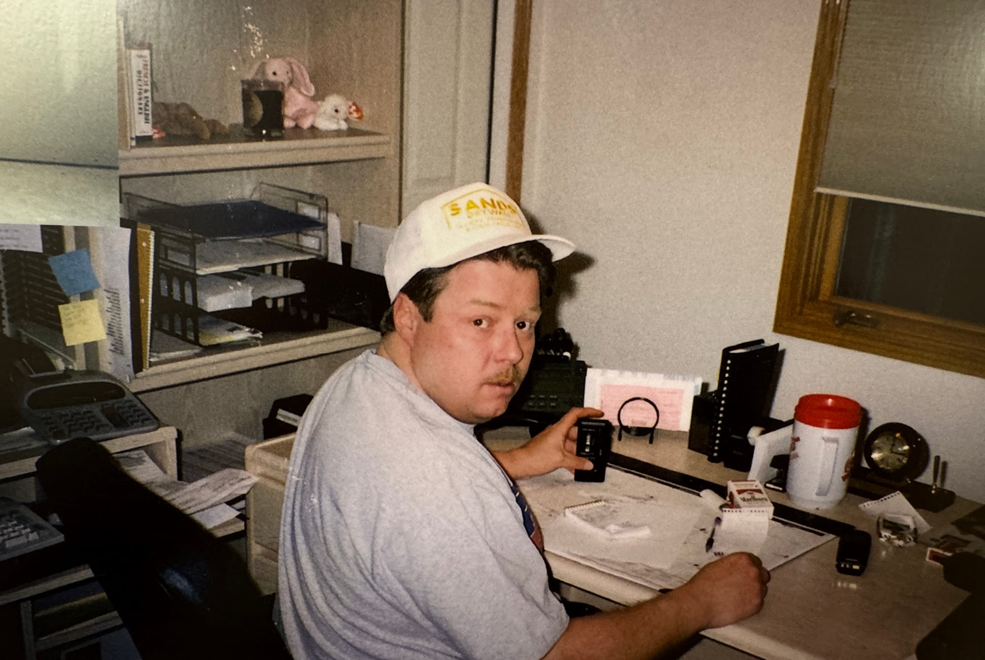 Greg Sands sits at a desk while wearing a baseball hat in the 1990s