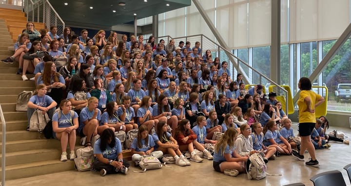 Dozens of girls sit on the stairs wearing blue shirts and are looking at a woman speaking in front of them.