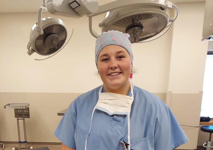 A female surgical tech wearing blue scrubs poses for a photo in a surgical room.